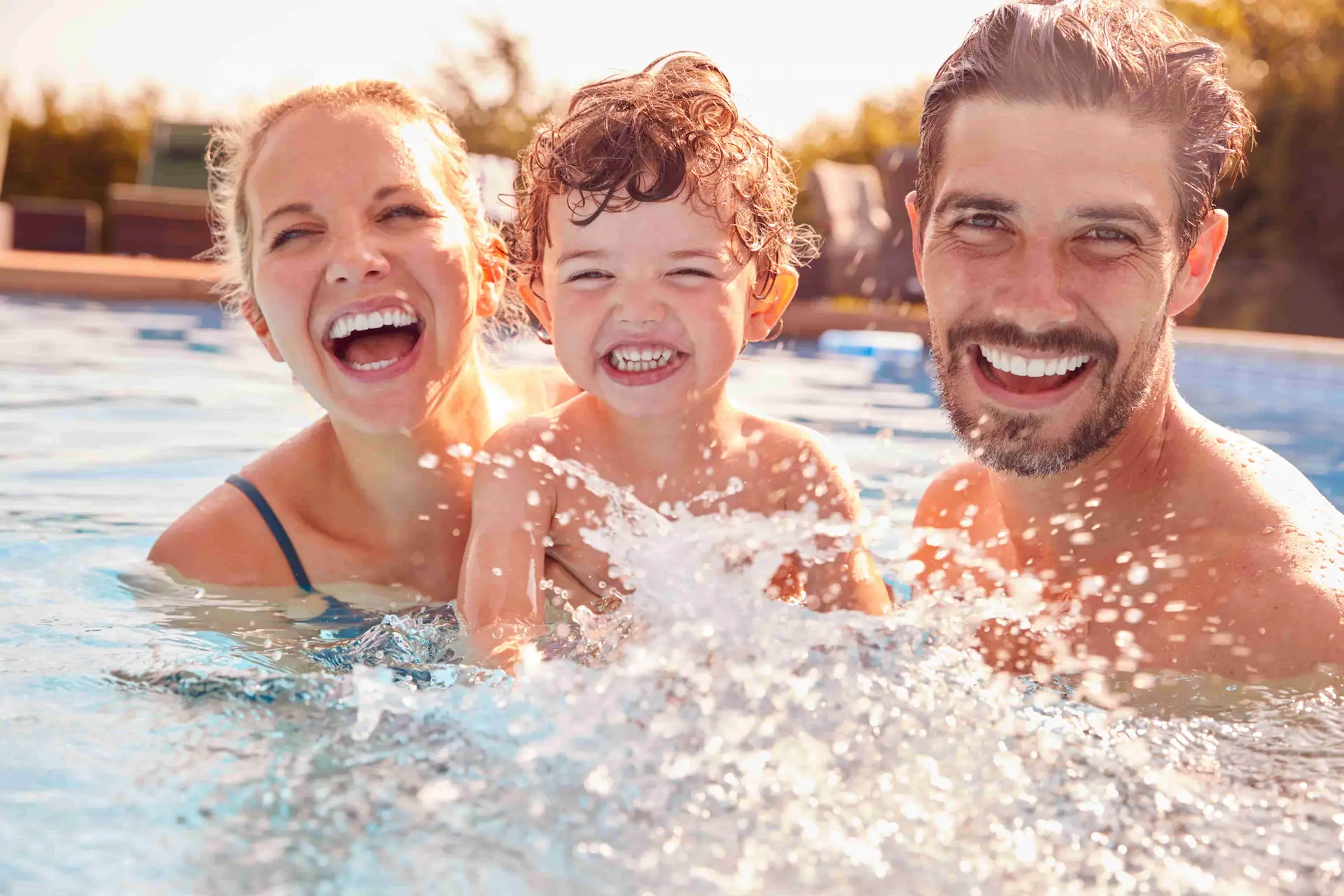 A man and woman splashing in the pool holding their toddler and smiling.