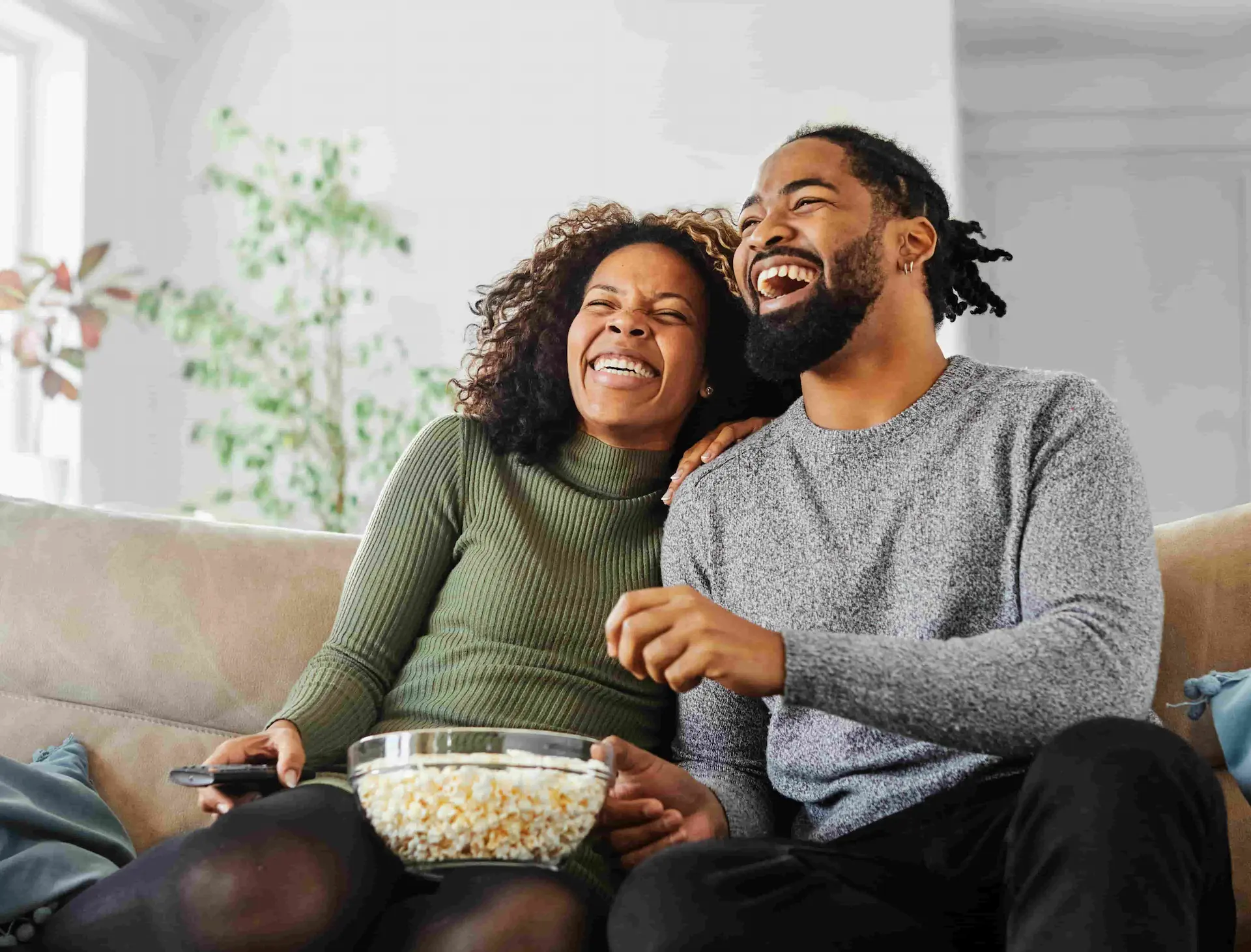 A black couple wearing sweaters sitting on a suede couch with a bowl of popcorn laughing out loud.